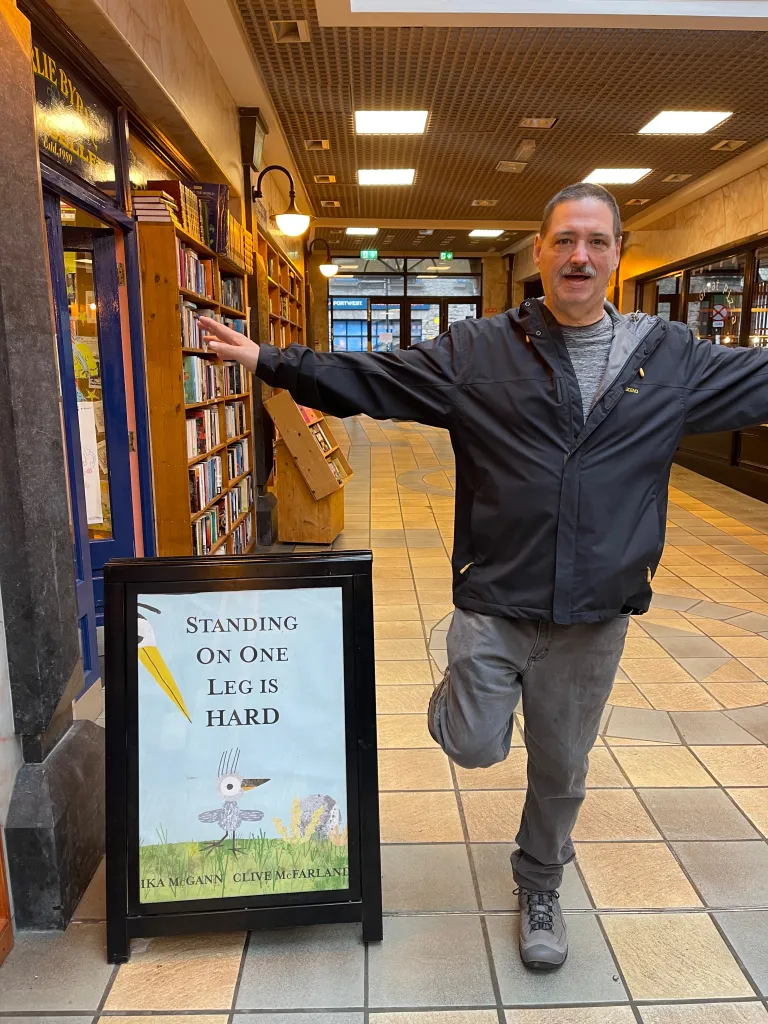 A man balances on one leg next to a sign that reads "STANDING ON ONE LEG IS HARD."