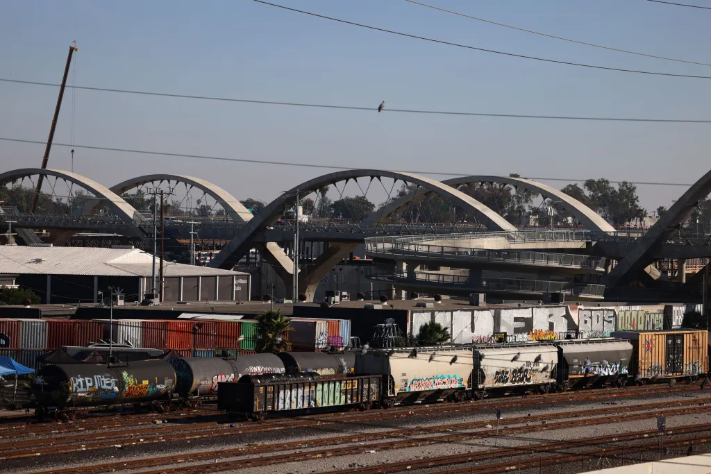 The 6th Street ribbon of light bridge in Los Angeles, spanning the Los Angeles River, with graffiti-covered trains and cargo containers below.