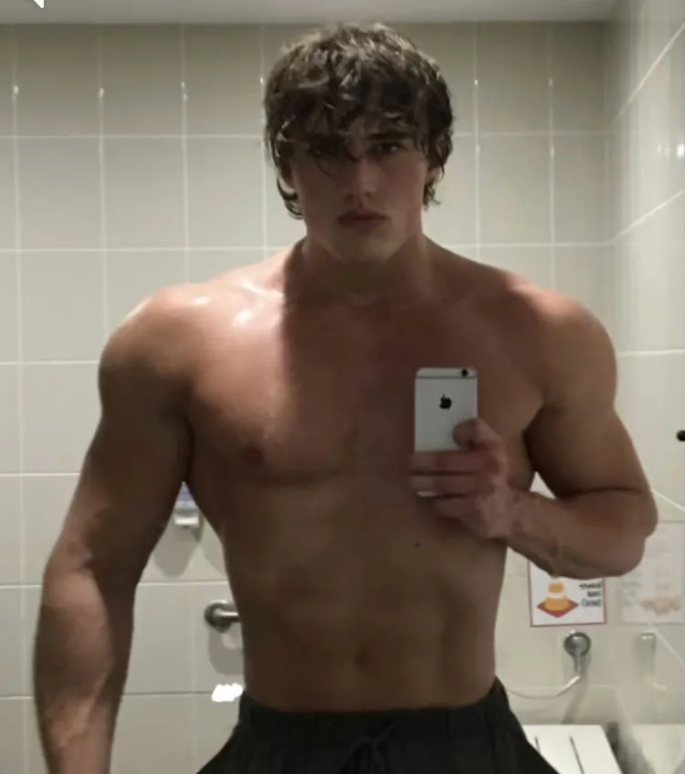 A shirtless man with short, wavy hair takes a mirror selfie in a tiled room, holding up a white iPhone.