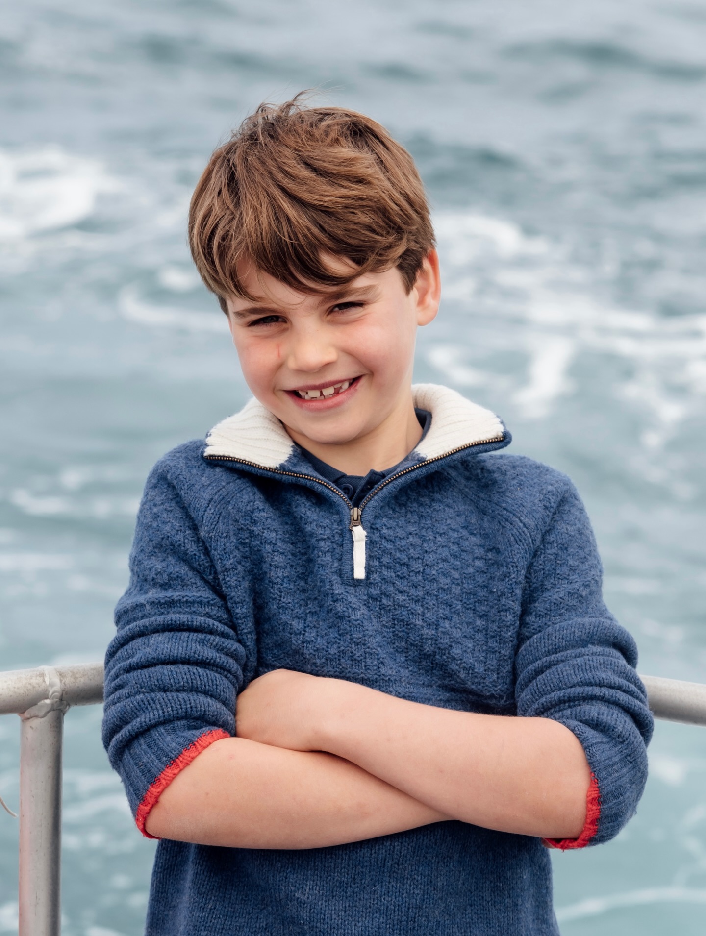 Prince Louis smiling in a blue sweater with arms crossed, with water in the background.