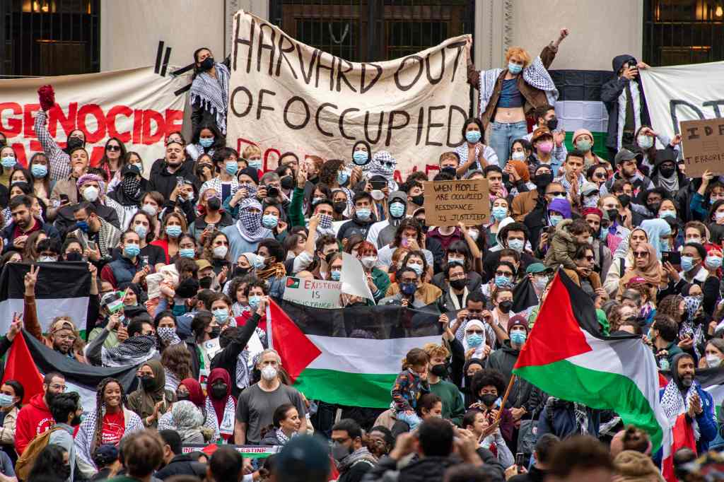 A large crowd of protesters holding signs, Palestinian flags, and a banner reading "HARVARD OUT OF OCCUPIED."