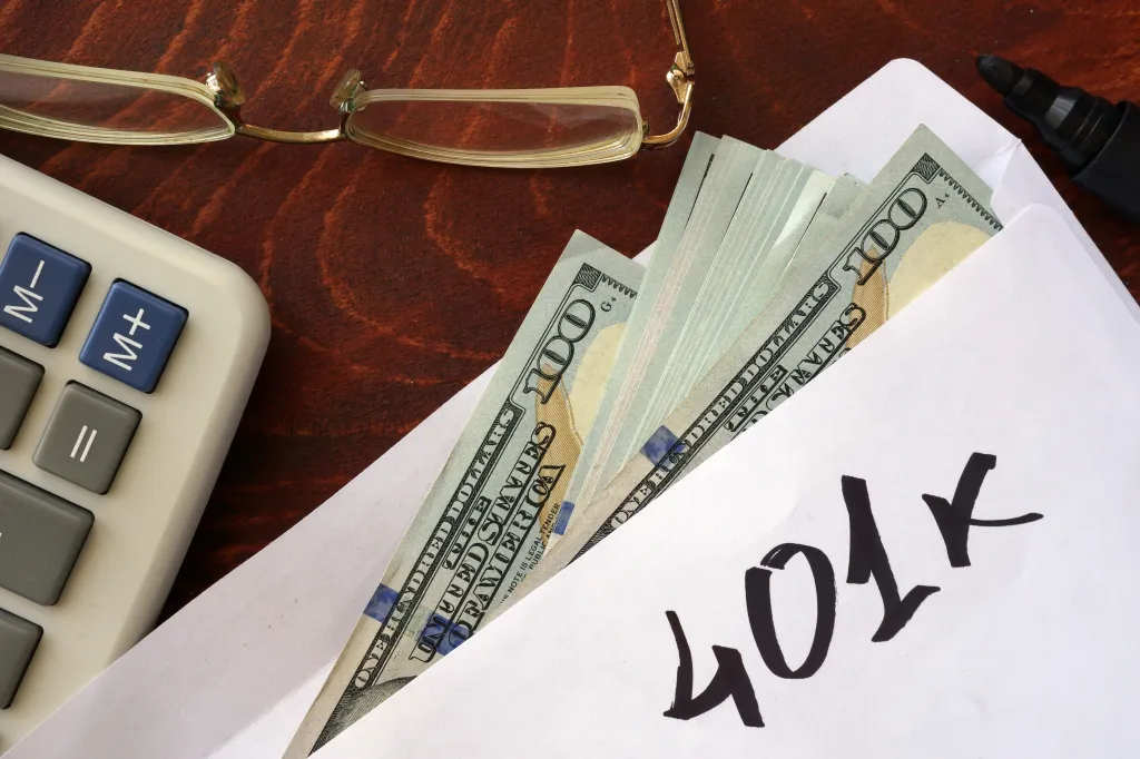 An envelope with "401k" written on it, filled with US dollar bills, next to a calculator and eyeglasses.