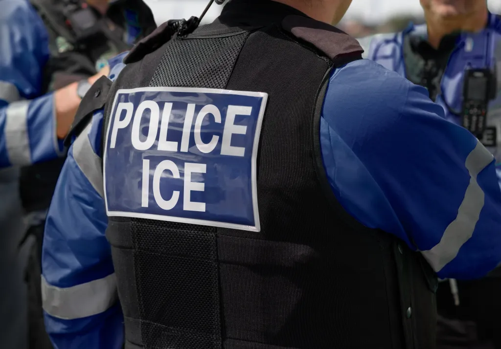 Rear view of an ICE Police agent in a black stab-proof vest with "POLICE ICE" written in white on a blue patch.