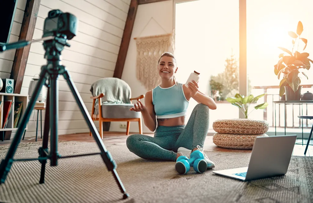 Woman blogger sitting on the floor with dumbbells and a laptop, showing a jar of sports nutrition proteins to the camera.