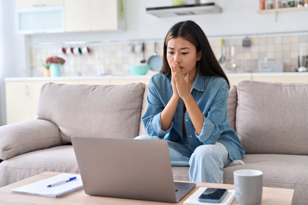 A nervous Asian woman looks at a laptop screen, worrying about bad news.