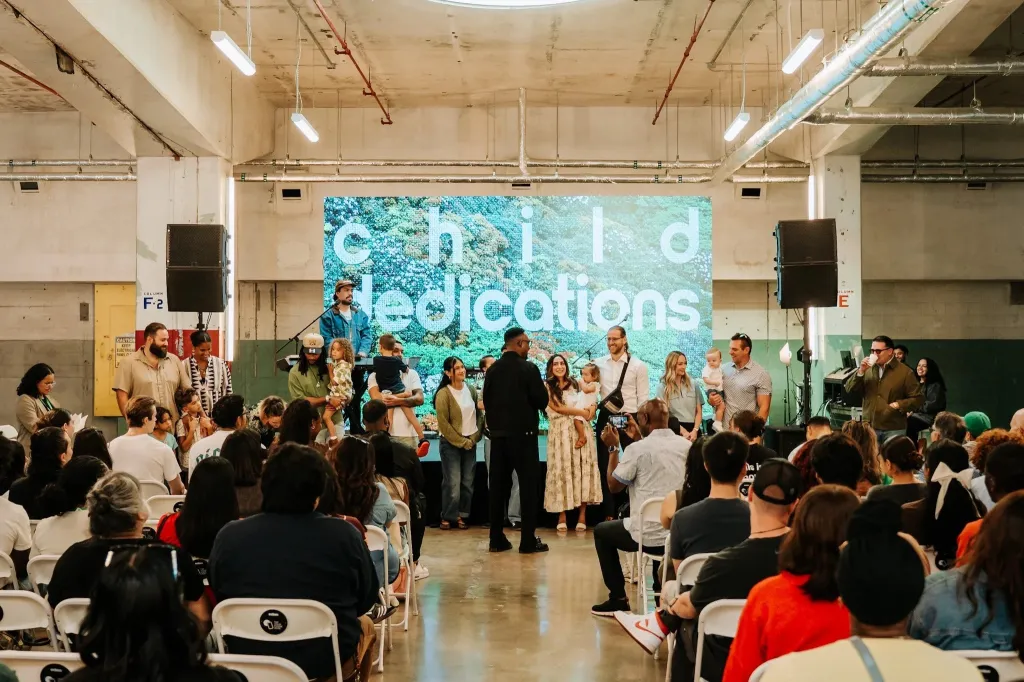 A group of adults and children on stage in front of a screen displaying "child dedications" to a seated audience.