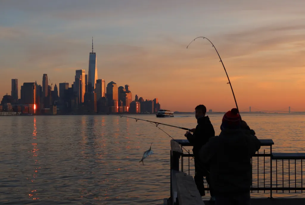 Two men fishing from a pier with a caught fish mid-air, with the New York City skyline and a ferry in the background at sunset.