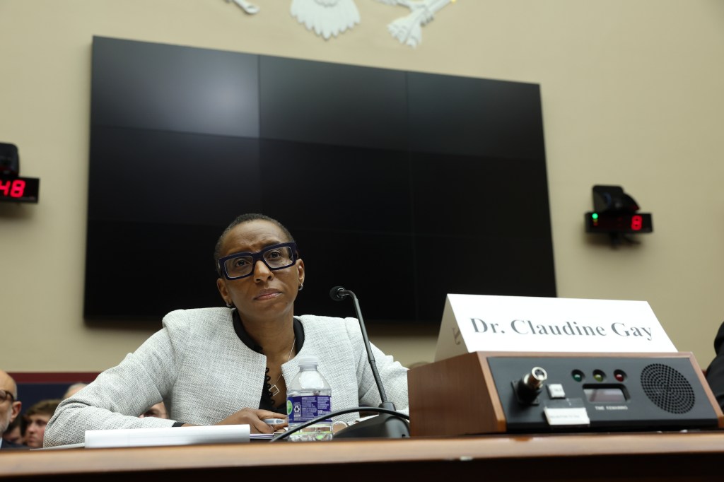 Claudine Gay, wearing glasses and a light grey jacket, sits at a table with a microphone and a "Dr. Claudine Gay" name card in front of her.