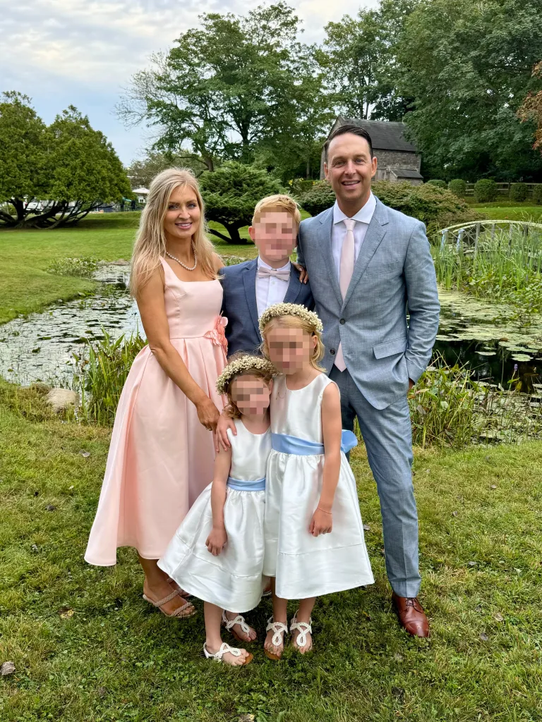 A family of five, including two adults and three children, posing in a garden with a pond and bridge in the background.