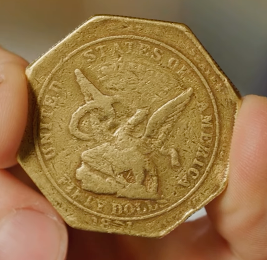A person holding an octagonal gold coin from 1851 with the words "UNITED STATES OF AMERICA" and "CALIFORNIA GOLD", with a depiction of an eagle and a Liberty head.
