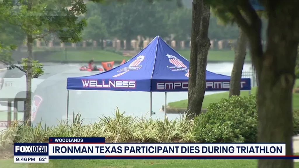 A blue "Wellness Empowerment" tent by a body of water with rescue boats, and a news banner stating "Ironman Texas Participant Dies During Triathlon."