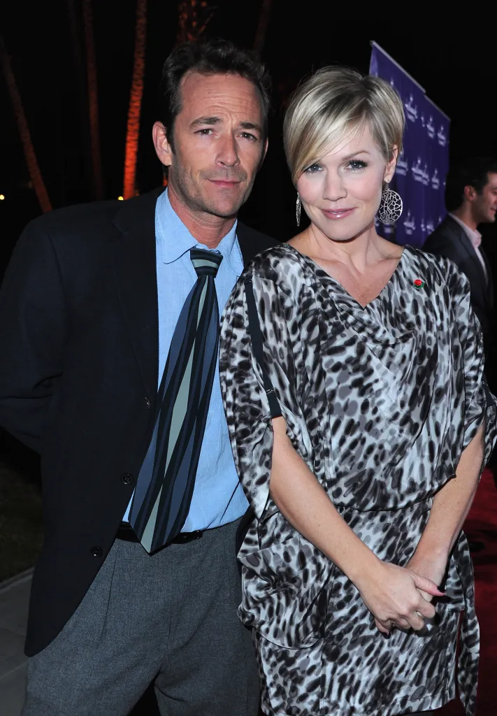 Luke Perry in a dark suit with a light blue shirt and striped tie, next to Jennie Garth in a black and white leopard print dress.