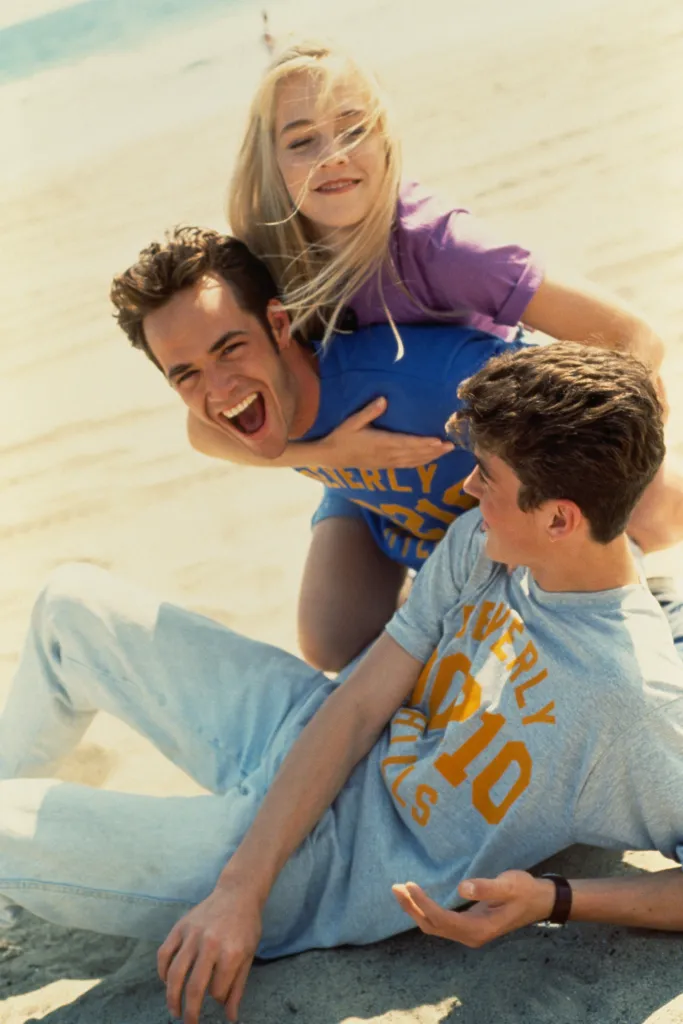 Jennie Garth on the back of Luke Perry, and Brian Austin Green lying on the sand, all wearing Beverly Hills, 90210 shirts.