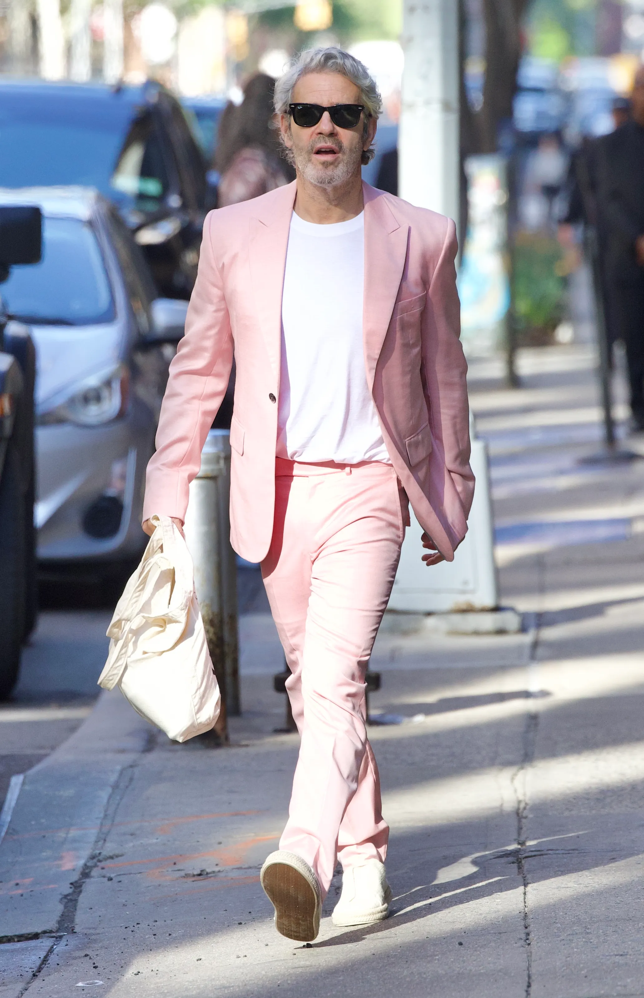 Andy Cohen walking down the street in a pink suit, white t-shirt, and white sneakers, holding a white bag.