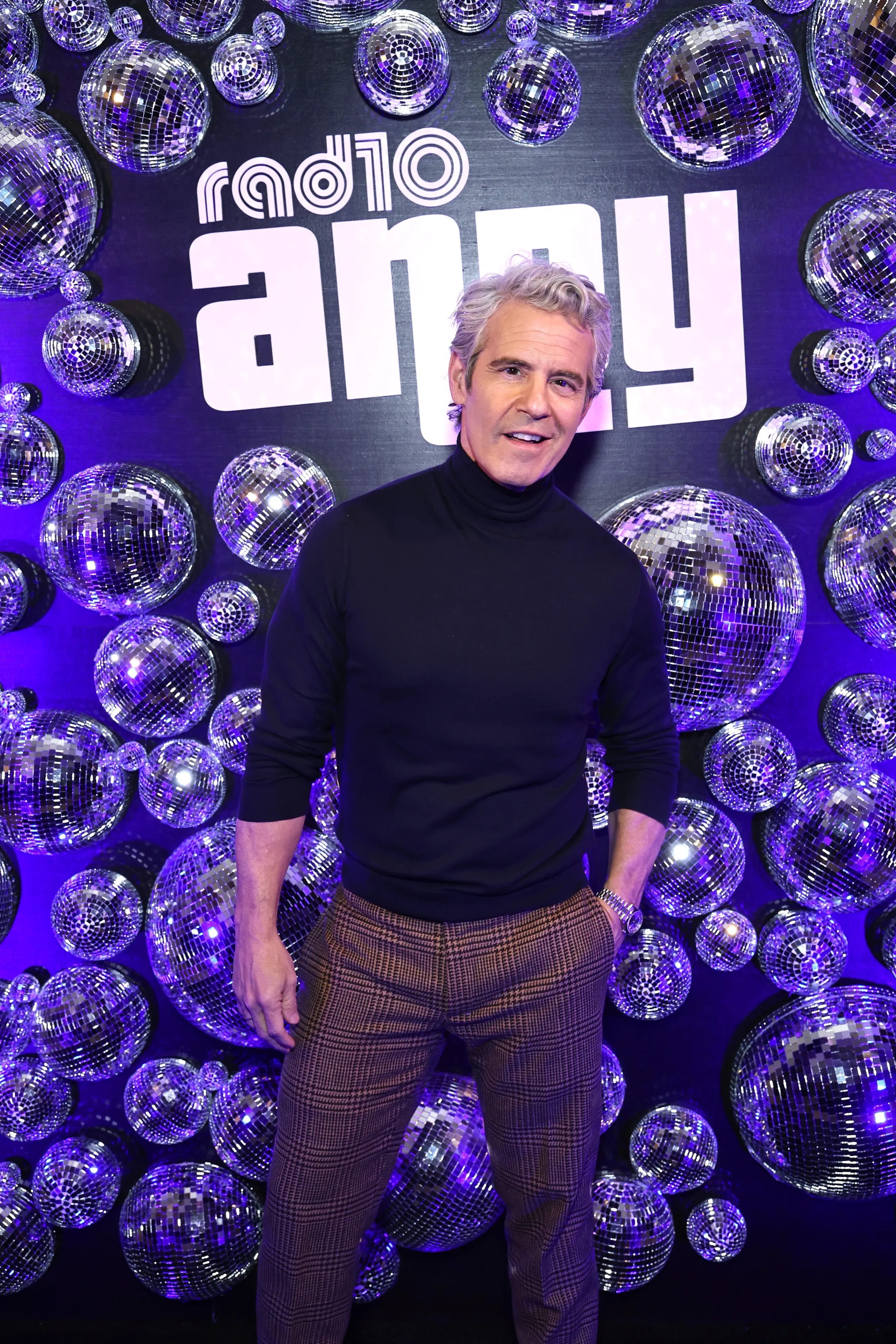 Andy Cohen in a black turtleneck and plaid pants, posing in front of a purple wall decorated with disco balls and the "Radio Andy" logo.