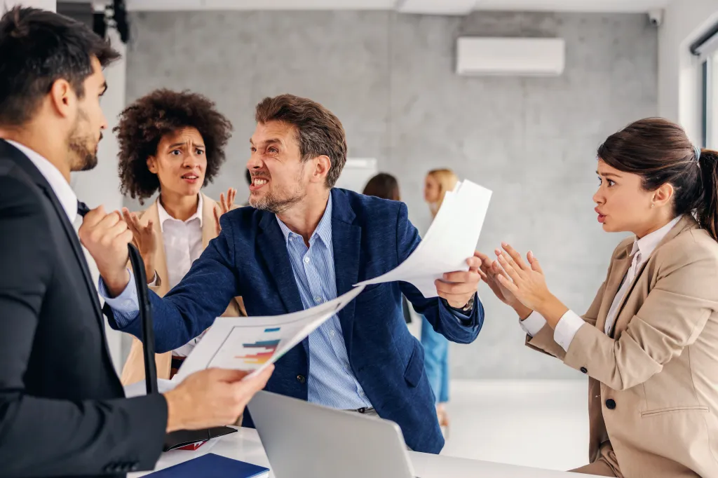 An angry boss holding his employee's tie and yelling while other coworkers look on in distress.