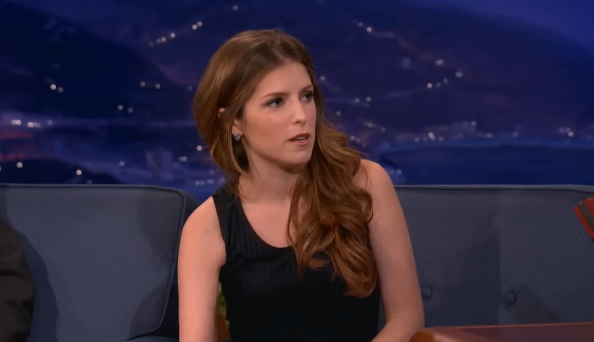Anna Kendrick on a blue couch, speaking during an interview.