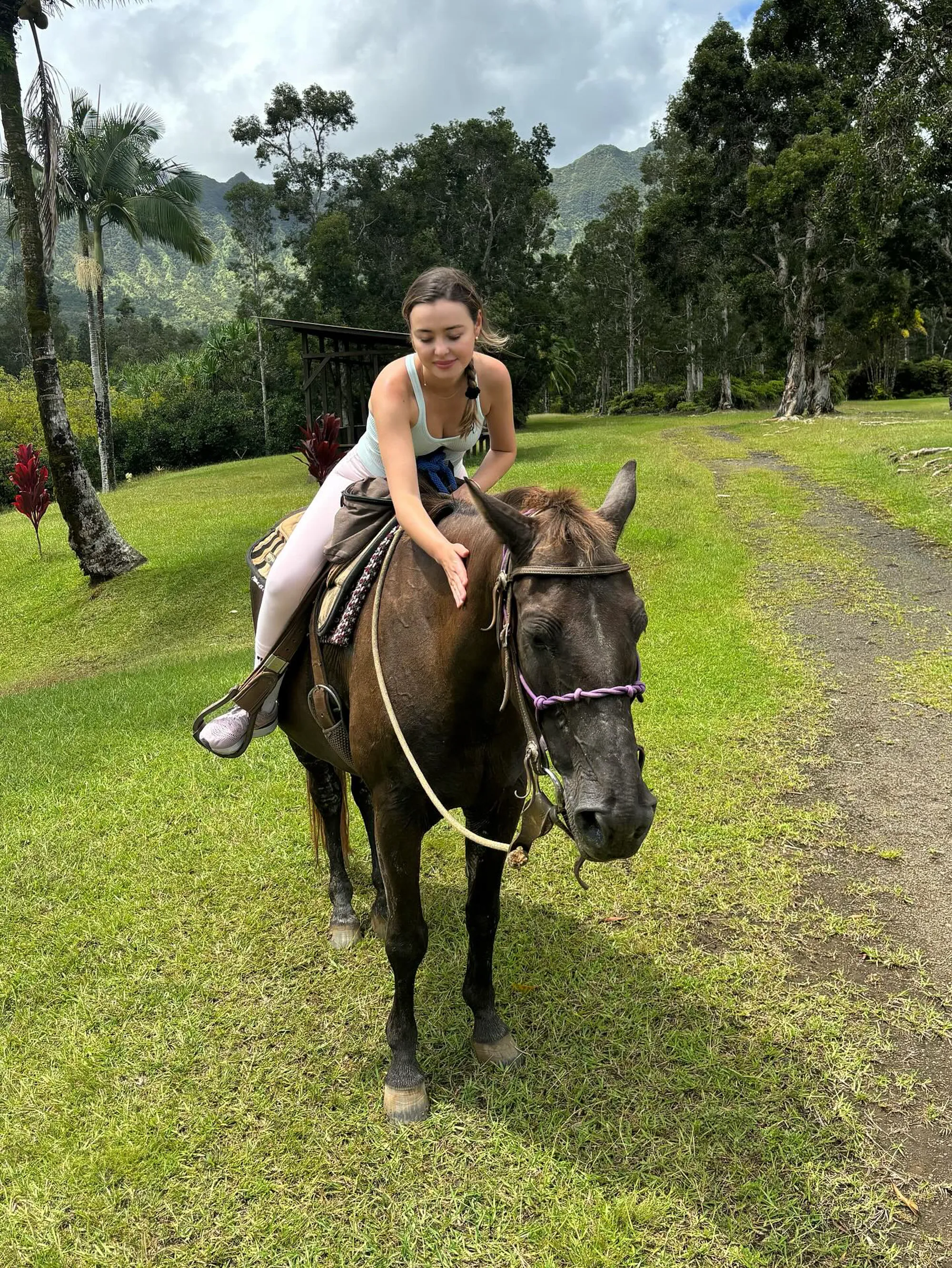 Eileen Kelly riding a horse, petting its neck, in a tropical landscape.