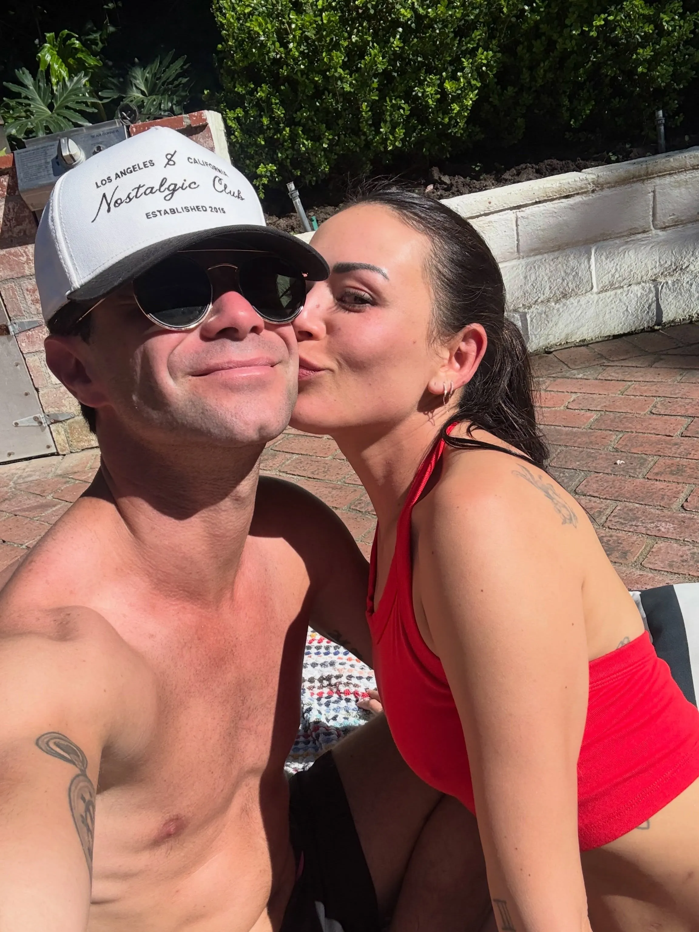 Sasha Farber in a white cap and Janel Parrish in a red top, with Parrish kissing Farber on the cheek.