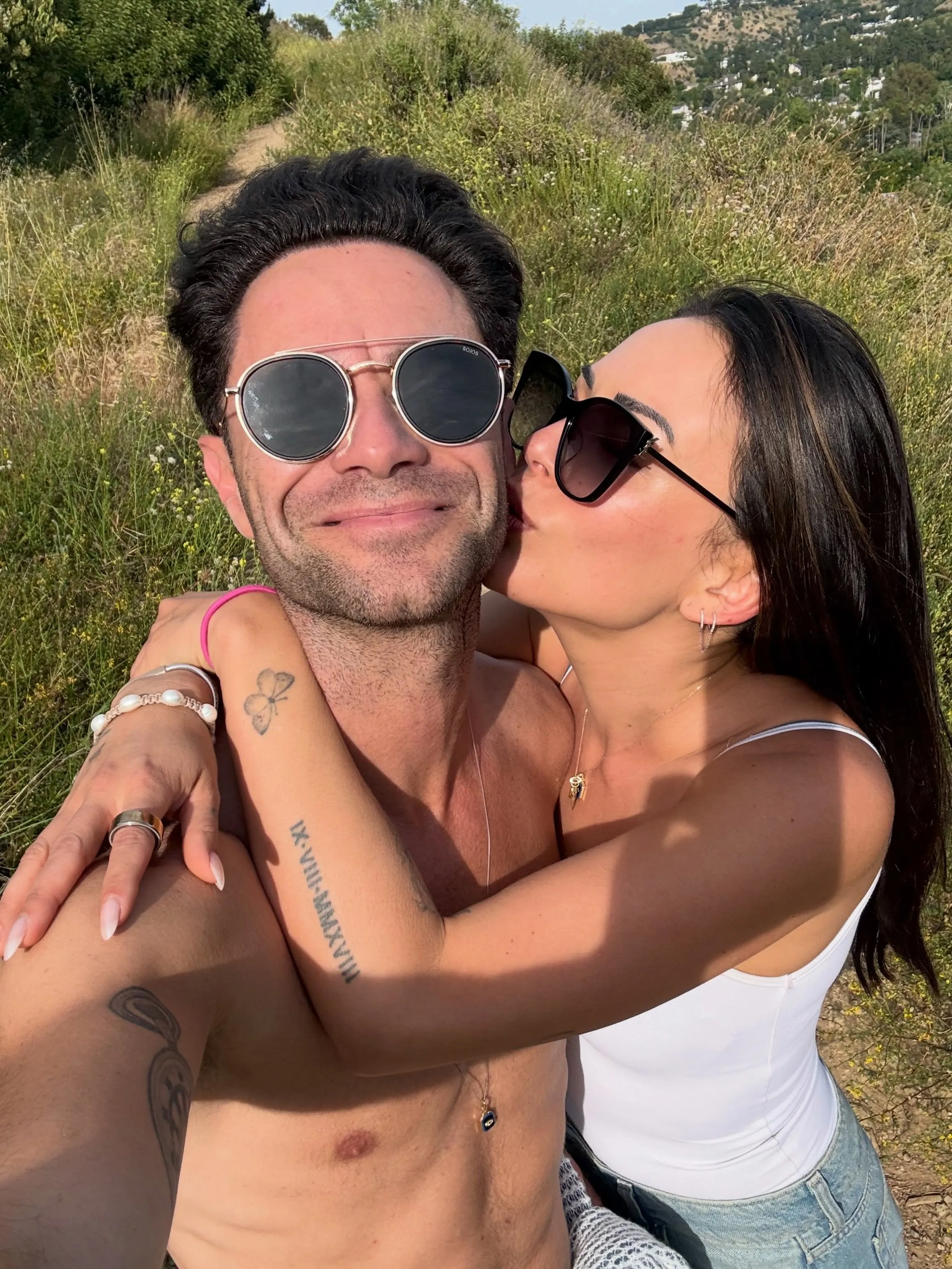Sasha Farber and Janel Parrish taking a selfie together in a field, with Parrish kissing Farber on the cheek.