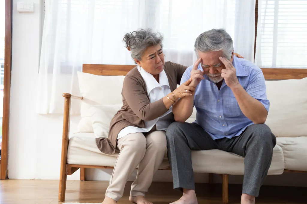 An elderly Asian woman comforts an elderly Asian man holding his head, likely due to dementia or Alzheimer's.
