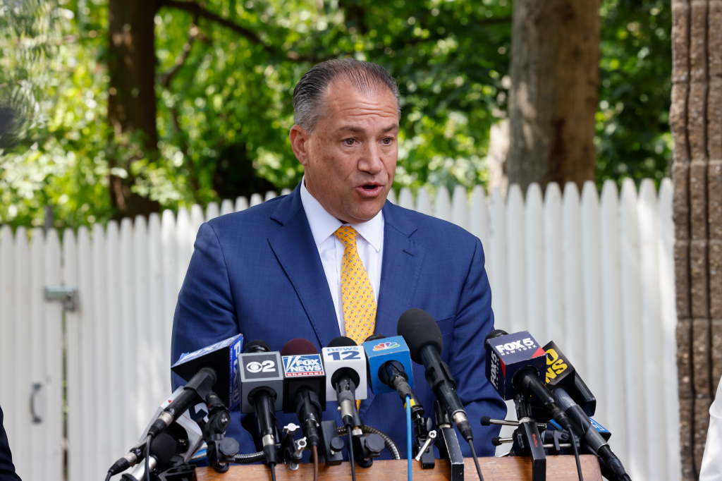Attorney Robert Macedonio speaking at a press conference surrounded by microphones from various news outlets.