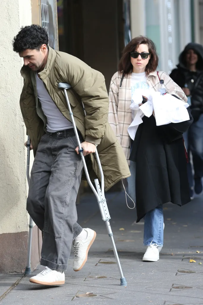 Christopher Abbott on crutches and Aubrey Plaza walk down a street.