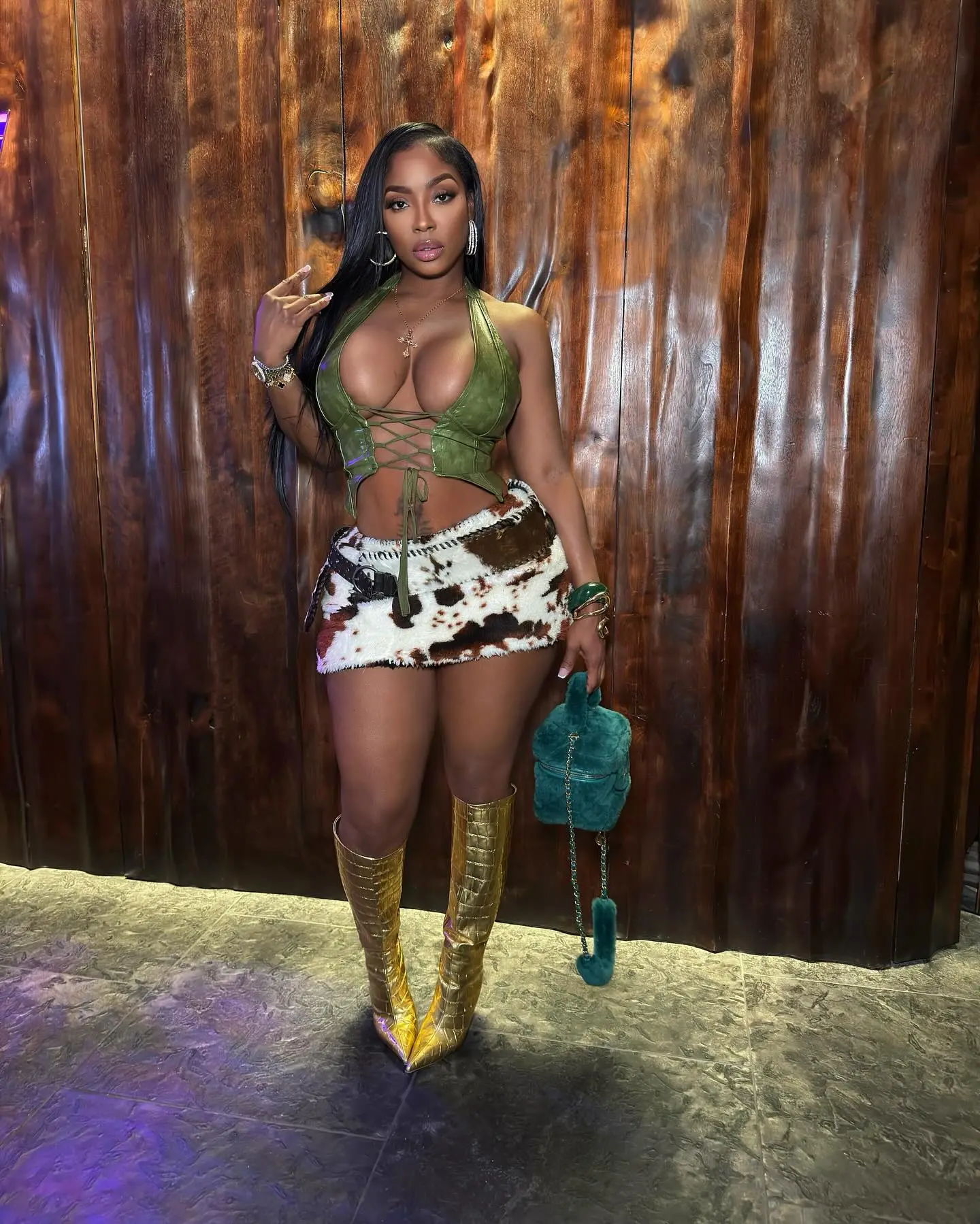 Ayesha Howard posing in a green lace-up halter top, a cow print mini skirt, and gold knee-high boots.