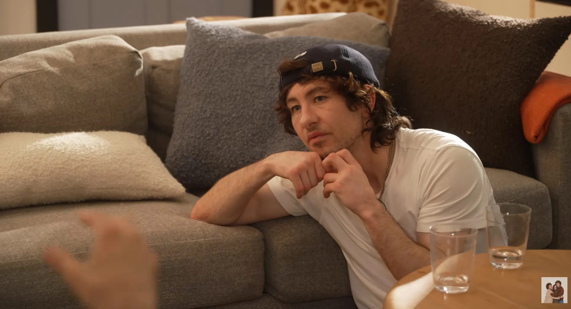 Barry Keoghan sits on the floor leaning against a gray couch, wearing a white t-shirt and a dark baseball cap.