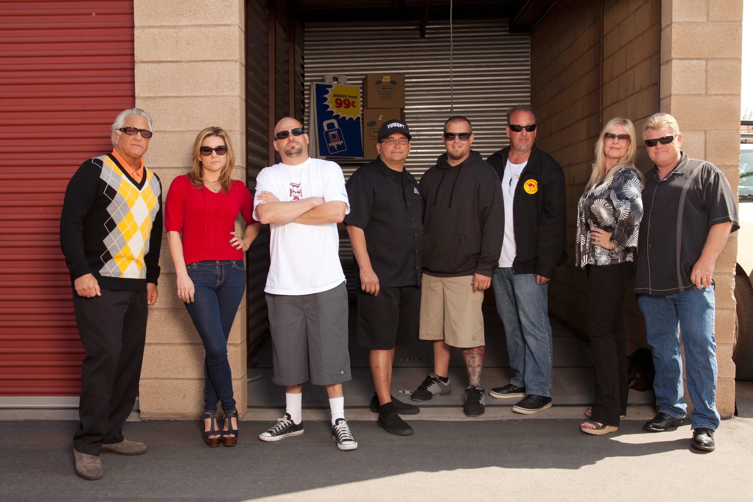 The cast of Storage Wars, Barry Weiss, Brandi Passante, Jarrod Schulz, Dave Hester, Brandon Sheets, Darrell Sheets, Laura Dotson, and Dan Dotson, stand in front of storage units.