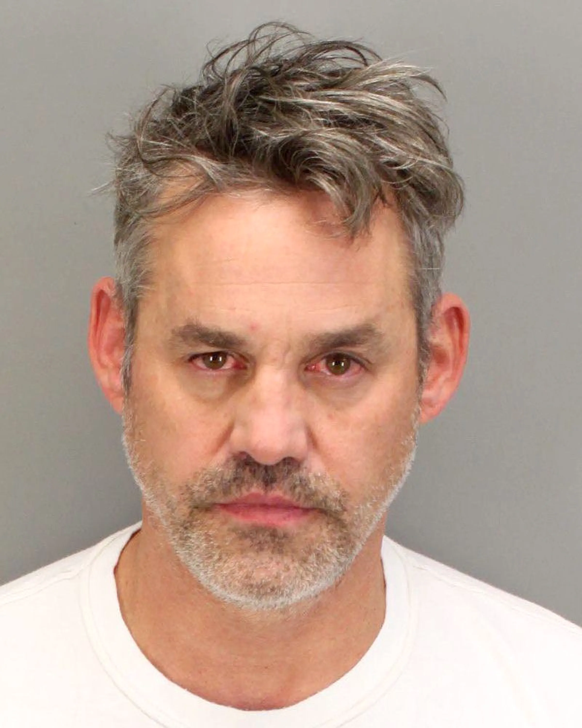 A close-up mugshot of Nicholas Brendon with red eyes, graying hair, and a beard.