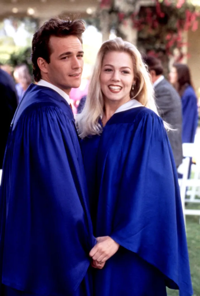 Luke Perry and Jennie Garth in cap and gown, holding hands.