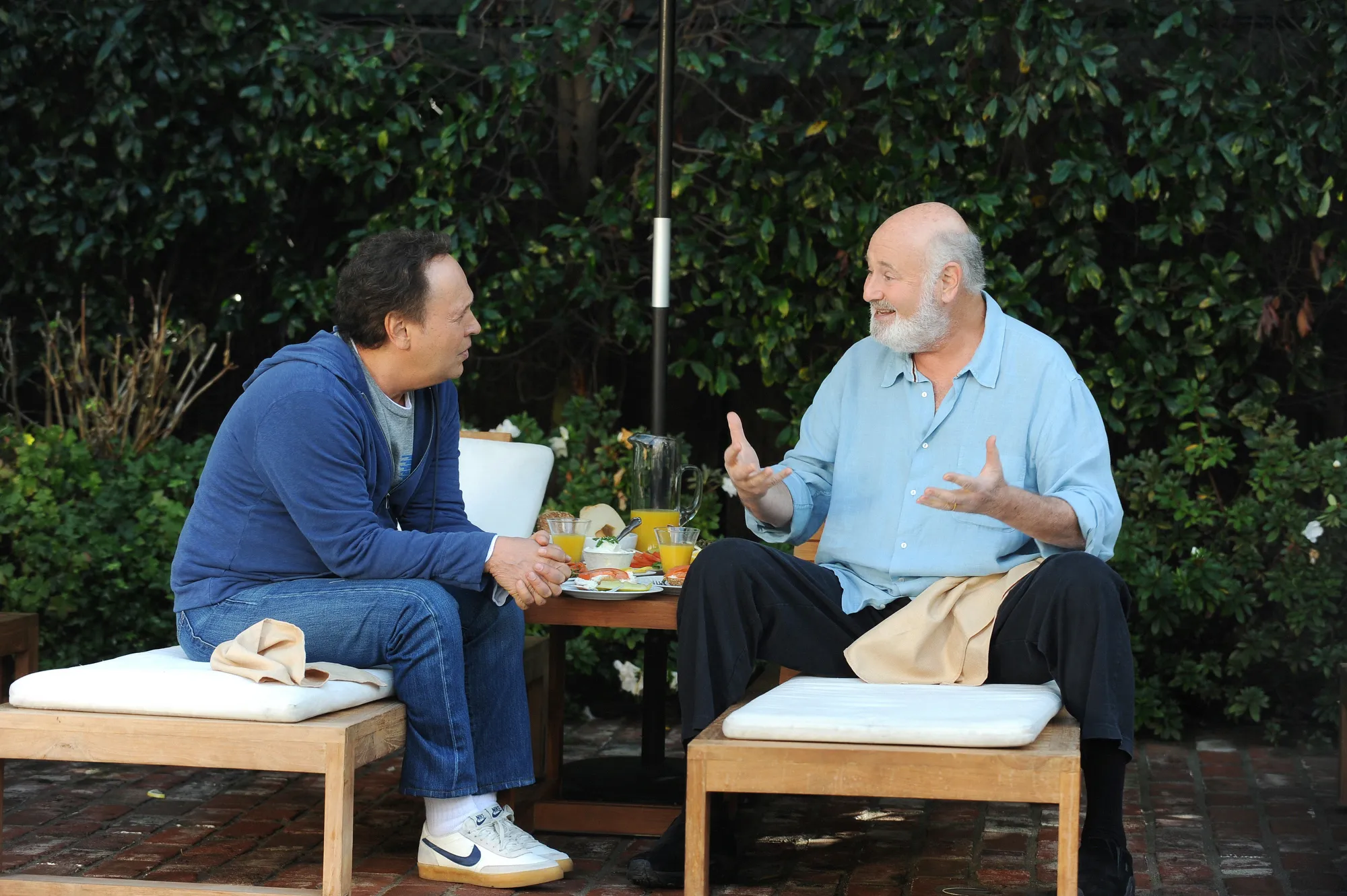 Billy Crystal and Rob Reiner talking outdoors, sitting on lounge chairs with breakfast on a table between them.