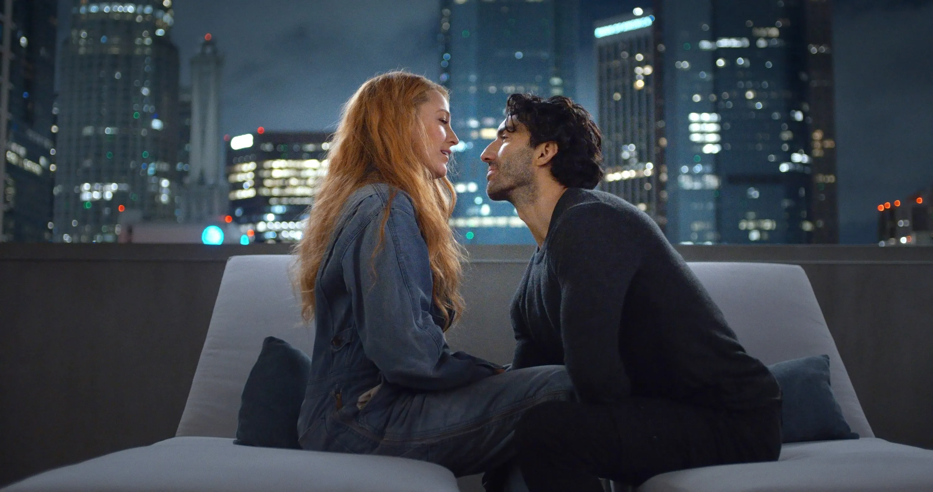 Blake Lively and Justin Baldoni looking at each other on a couch with a city skyline at night.