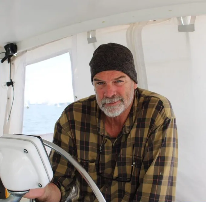 Brian Hooker, a man with a gray beard and a dark beanie, smiles while steering a boat.