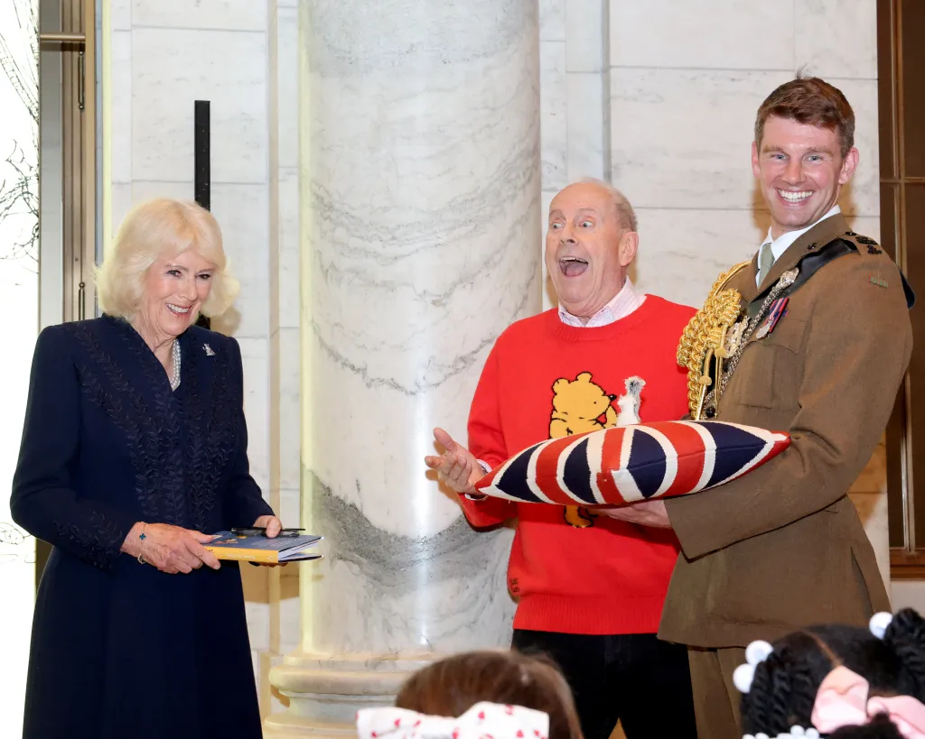 Queen Camilla, in a navy dress, holds a copy of "Winnie the Pooh" while a Gyles Brandreth n a red Winnie the Pooh sweater gestures animatedly next to a man in a military uniform holding a Union Jack pillow.