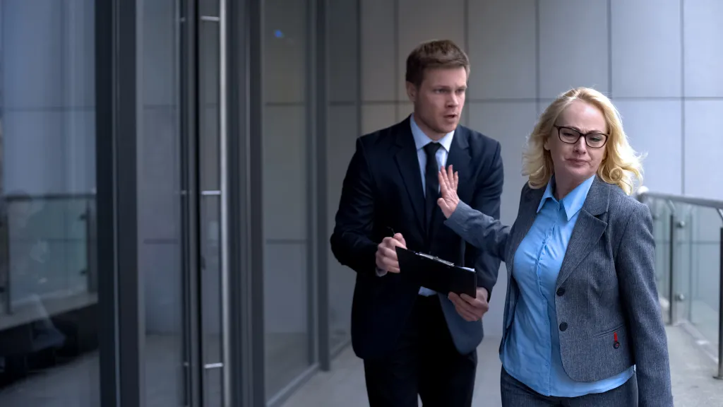 A businesswoman walking away from a young businessman who holds out a report on a clipboard to her.