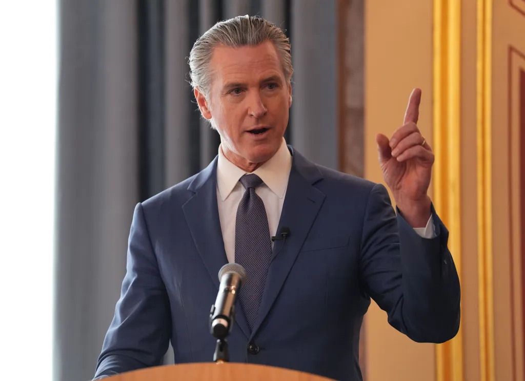 California Governor Gavin Newsom speaking at a podium with his hand raised.