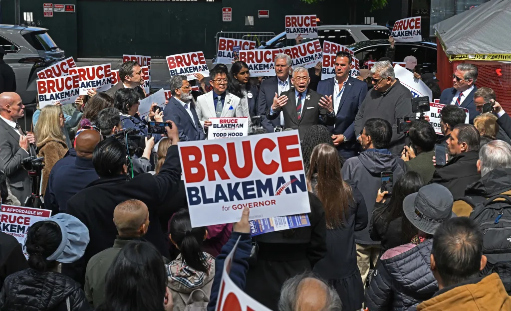 Bruce Blakeman speaking at a protest against a homeless shelter, with attendees holding signs for "Bruce Blakeman for Governor."