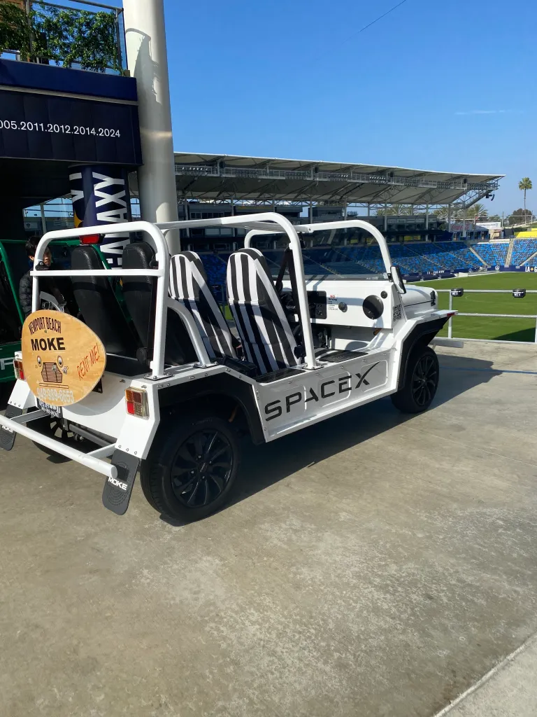 A white Moke electric vehicle with black and white striped seats, branded with "SpaceX," parked next to a stadium.