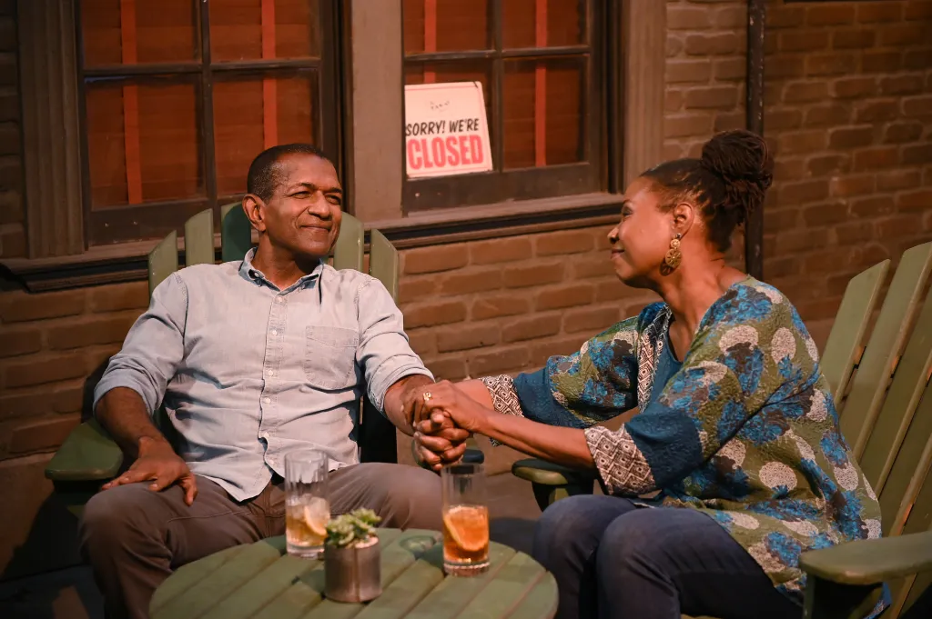 RIF HUTTON and JOYCE GUY sit in green chairs holding hands, with drinks on a small table in front of them.