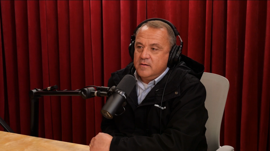 Chris Mikkelsen speaking into a microphone with headphones on, against a red curtain background.