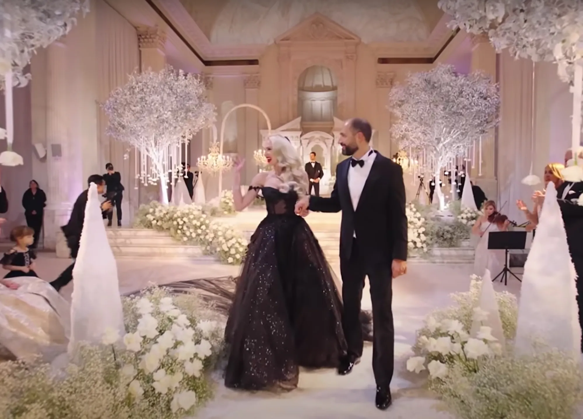 Christine Quinn and Christian Dumontet walk hand-in-hand down a white aisle lined with white flowers.