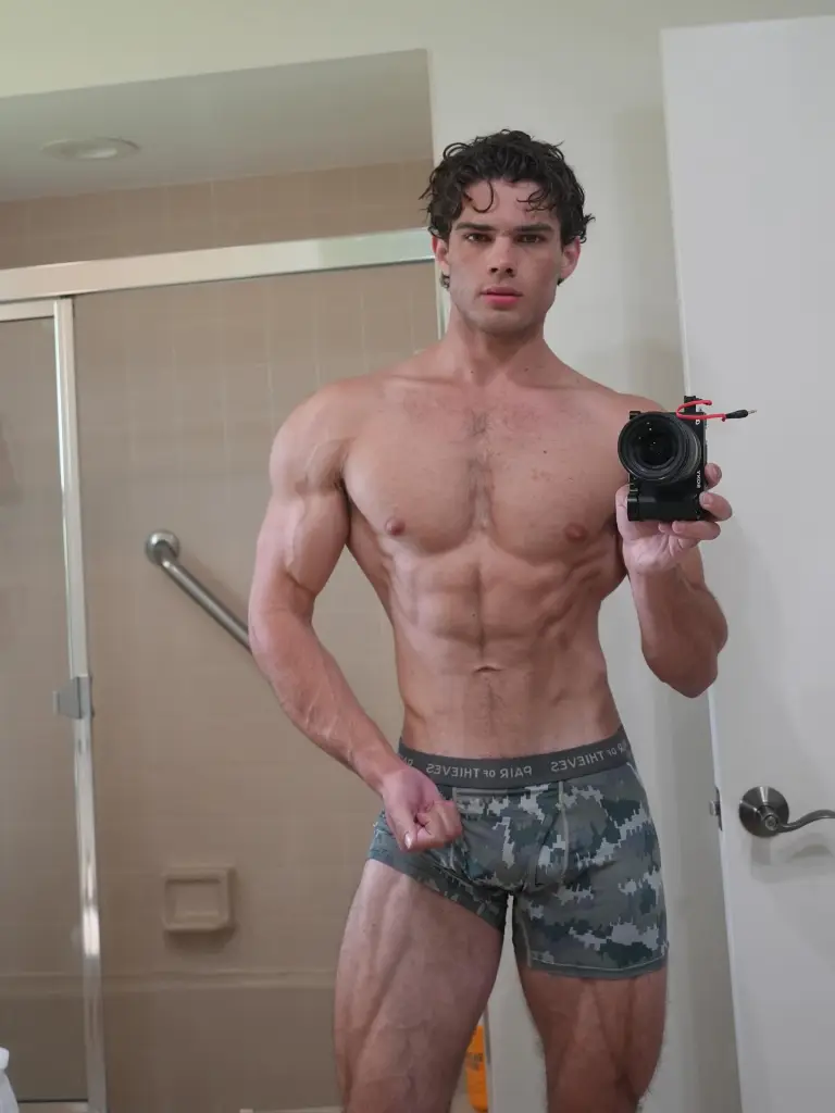 A shirtless man with a muscular physique takes a mirror selfie, holding a camera in his right hand.