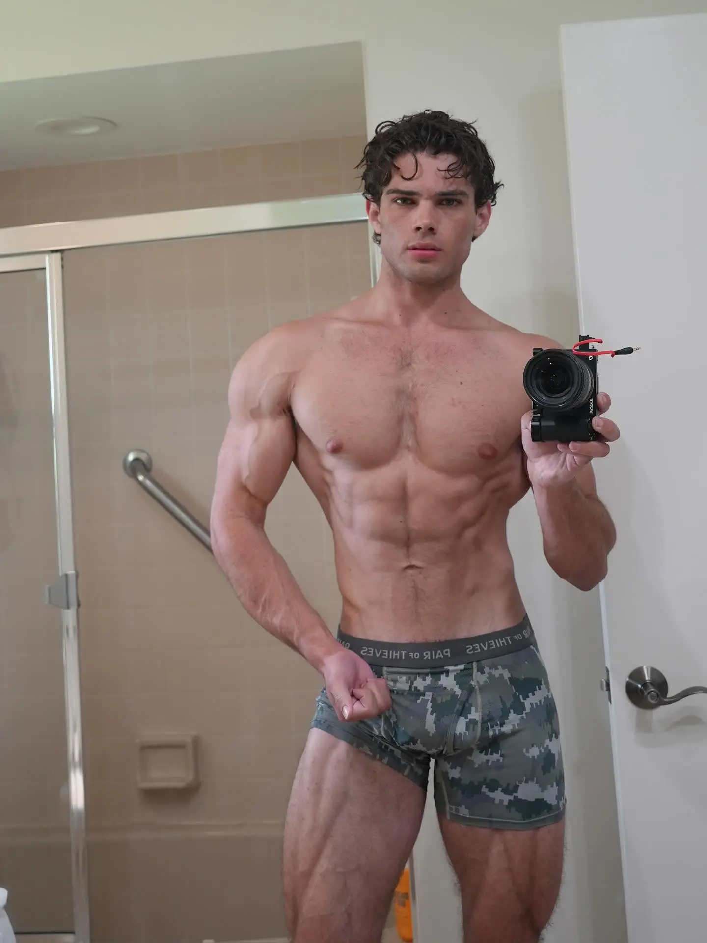 An influencer takes a selfie in a mirror, showing his sculpted physique while wearing camo boxer briefs.