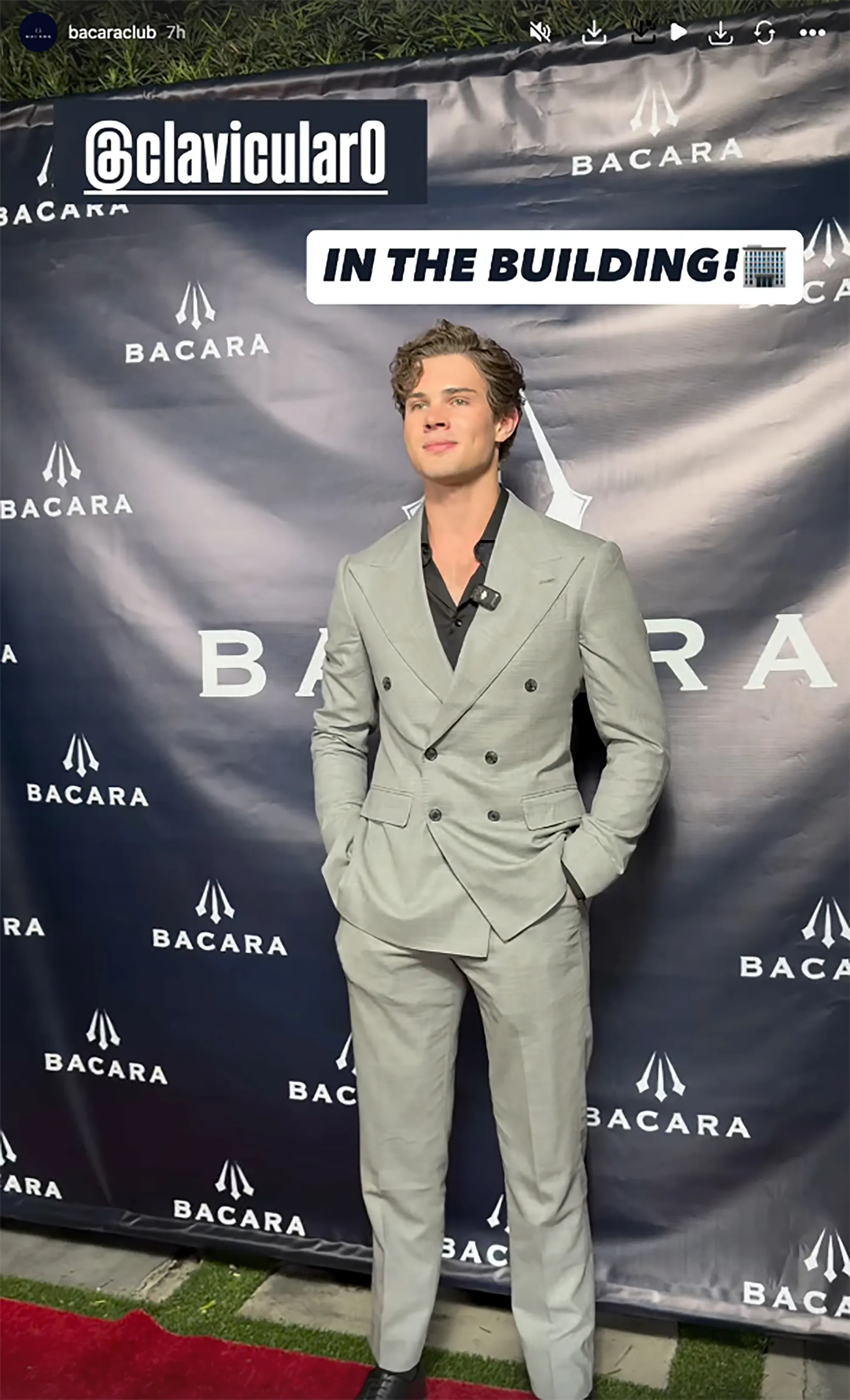 Clavicular in a light grey suit with a black shirt stands on a red carpet in front of a dark blue banner with the Bacara logo.