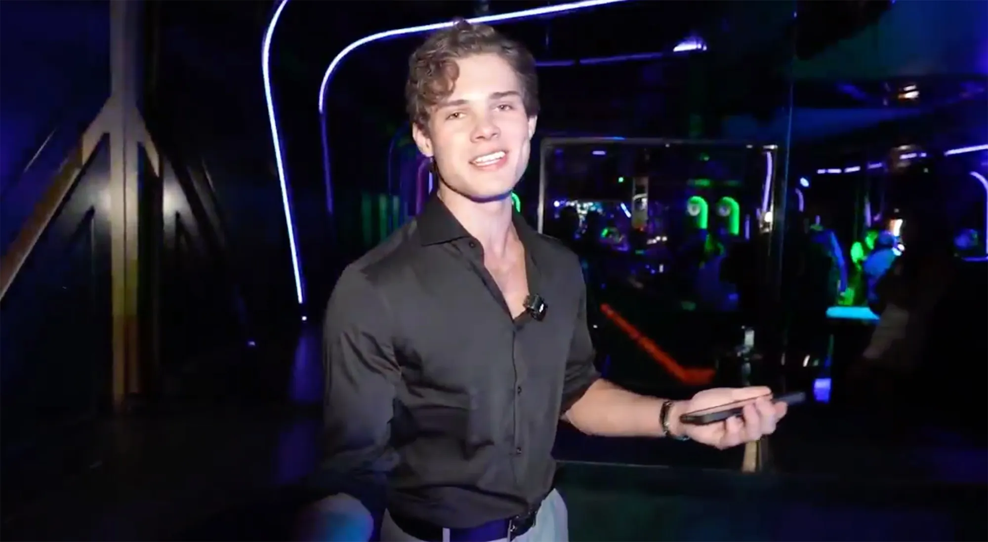 Clavicular in a black button-up shirt smiling while holding a phone in a dimly lit club with blue and green neon lights.