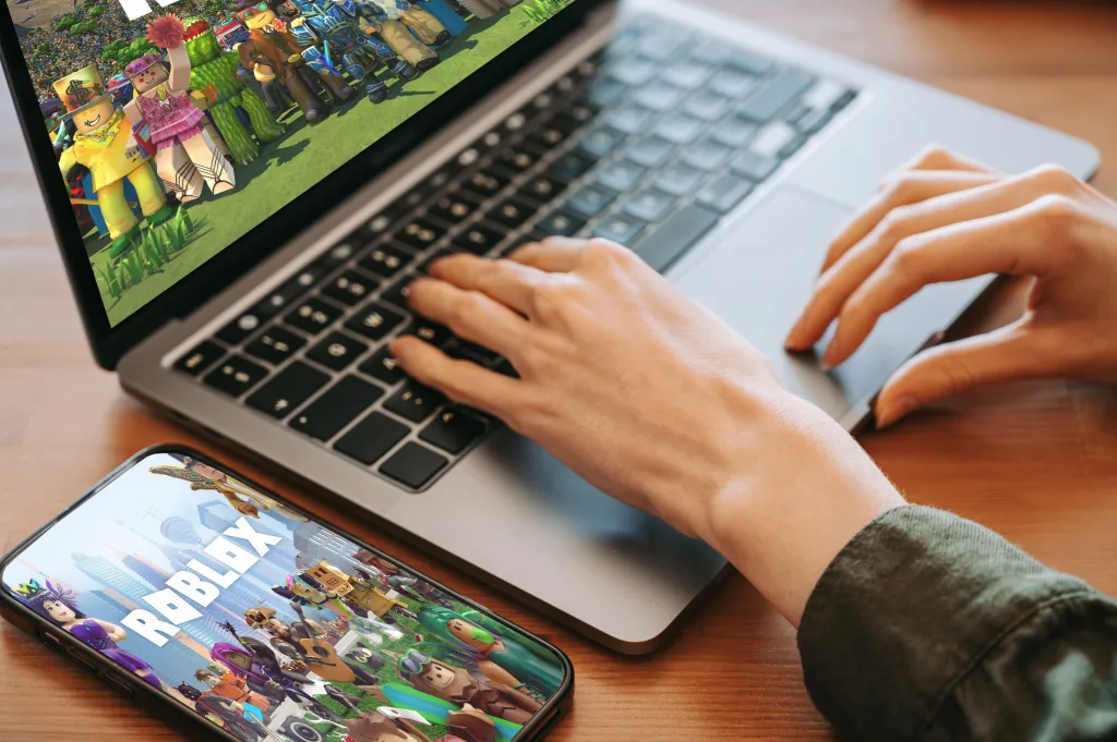 A person's hands typing on a laptop with the Roblox game on screen, and a smartphone displaying Roblox nearby.