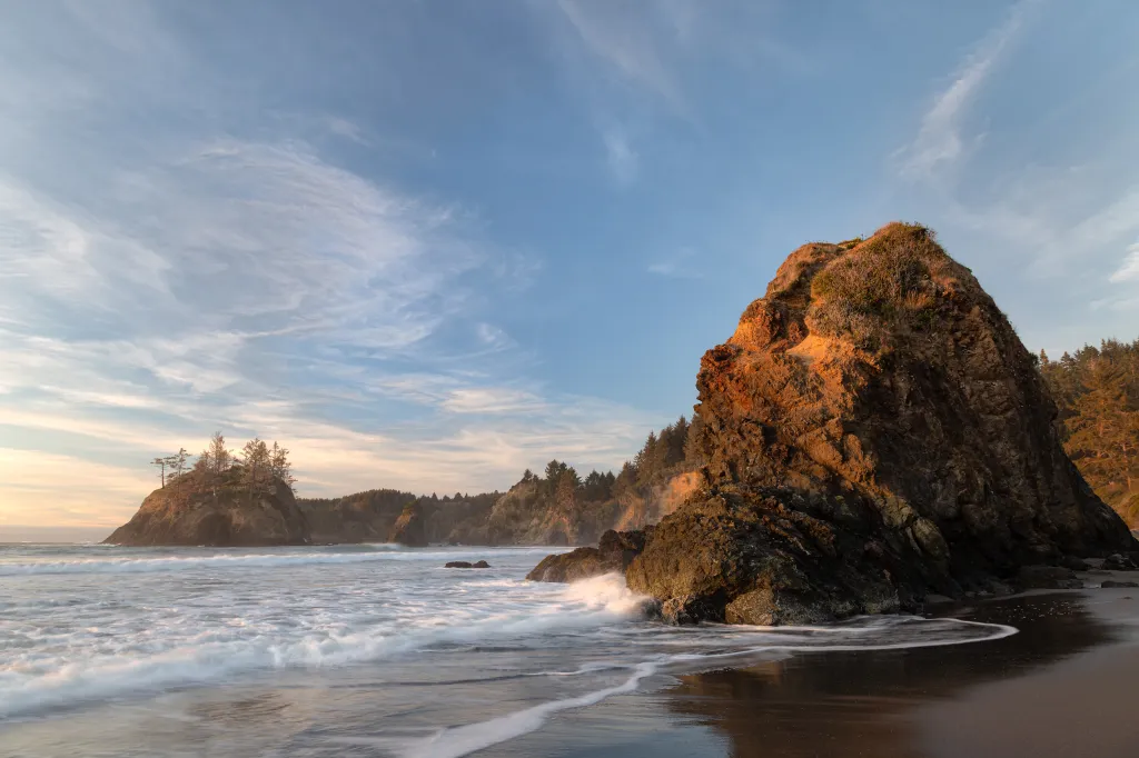 Seascape at sunset in Humboldt County, California, showing waves crashing on rocks and sandy beach, with tree-covered cliffs in the background.