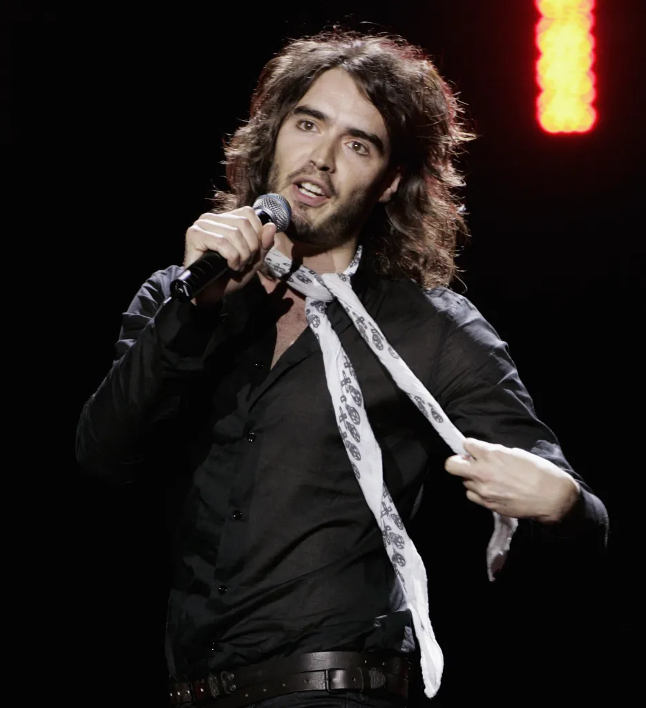 Russell Brand performing on stage, holding a microphone in one hand and his tie in the other.